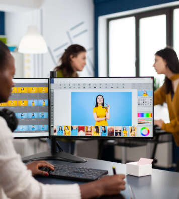 Black retoucher woman analysing images on pc with two monitors and retouching digital assets with stylus pencil. African project designer using photo editing software sitting in creative agency office