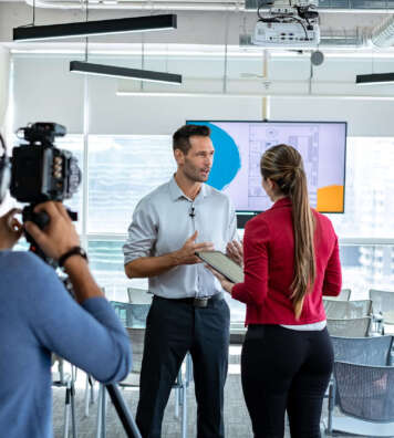 Happy businessman during corporate interview with female journalist. Manager answering question in office. Young woman at work as reporter with business man and cameraman shooting video for broadcast